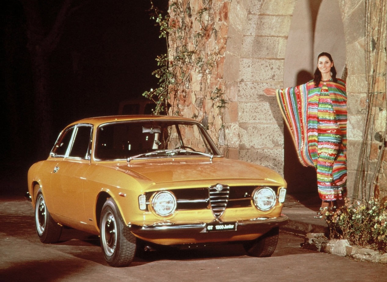 Voiture Alfa Romeo Giulia Coupé 1300 GT Junior modèle de 1966, de couleur marron clair métallisé, stationnée à côté d'une femme portant une longue robe colorée.