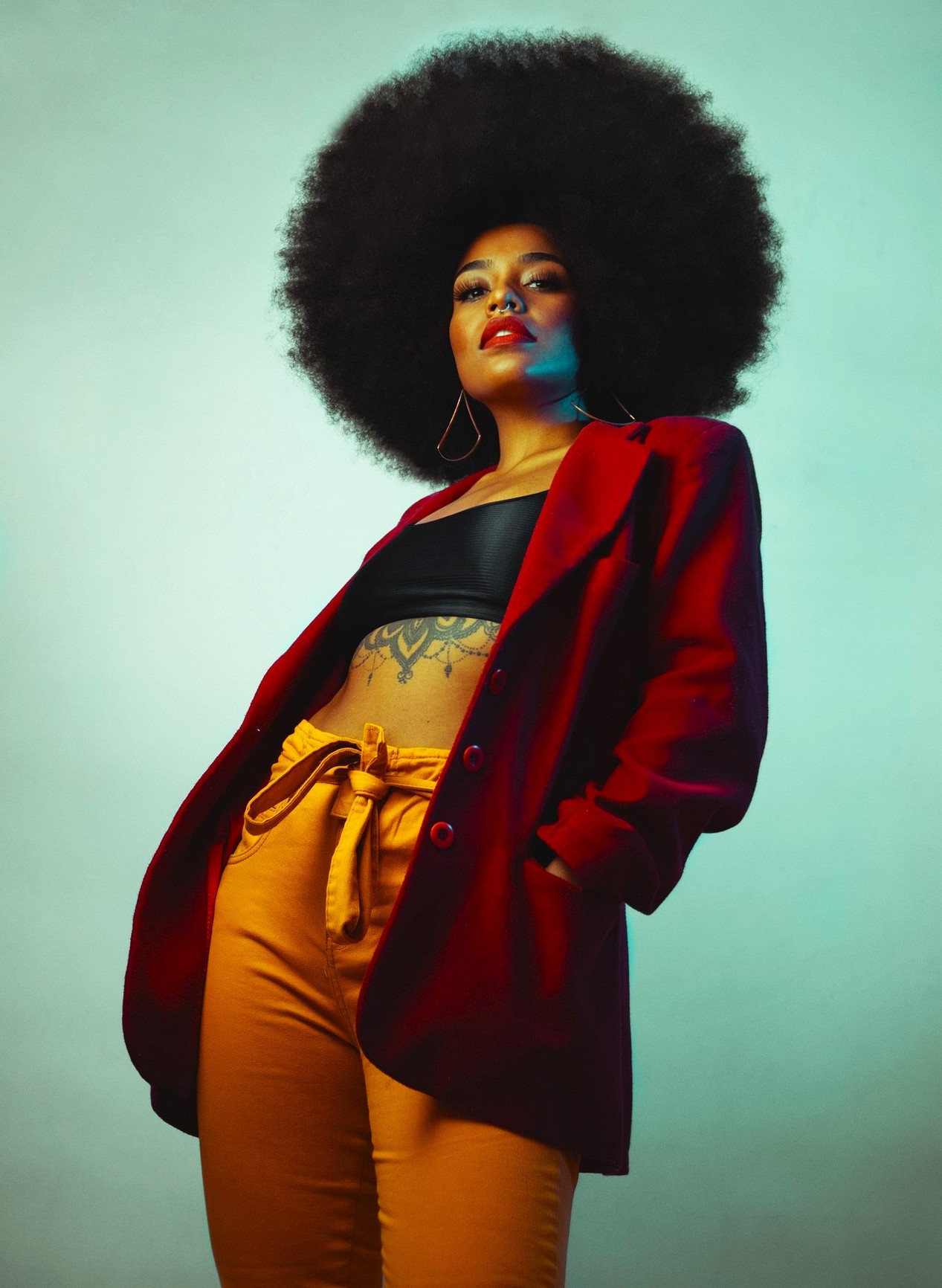 Belle femme noire avec une coupe afro, un top noir, une veste rouge et un pantalon orange.