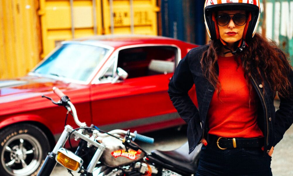 Femme portant les vêtements pour moto de la marque française Wildust Sisters, avec une moto noire et une auto rouge vintage