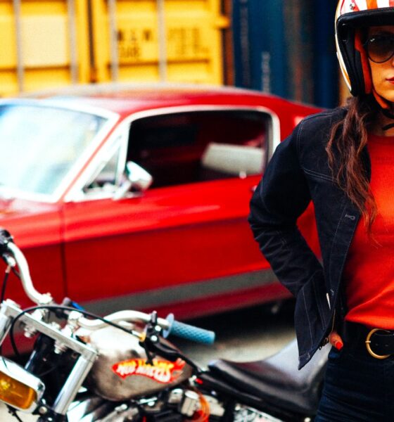 Femme portant les vêtements pour moto de la marque française Wildust Sisters, avec une moto noire et une auto rouge vintage